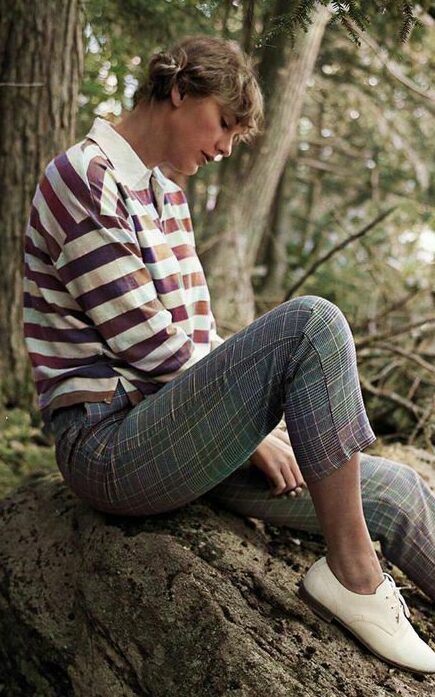 Taylor Swift sentada em uma floresta, usando uma camisa polo de manga longa e listras, calça skinny com estampa xadrez e sapato oxford.