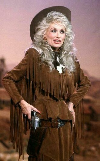 Dolly Parton vestida com roupas de camurça marrom e franjas, combinando com chapéu de cowboy.