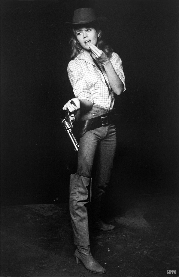 Jane Fonda no set de "Dívida de Sangue", usando botas, jeans, camisa xadrez e chapéu de cowboy.