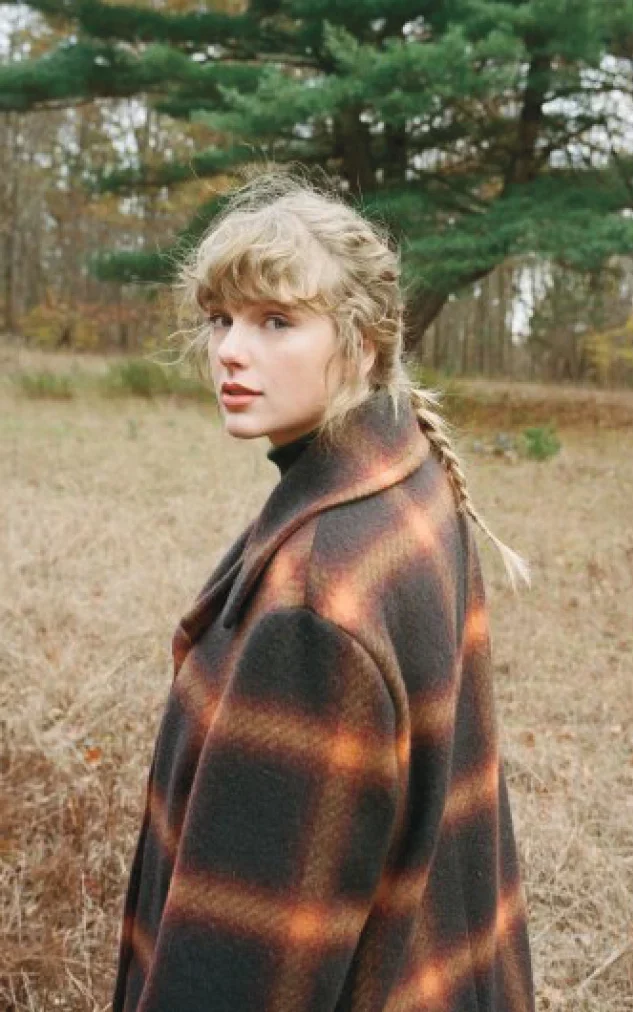 Taylor Swift posa em uma foleresta, vestindo um casaco longo e largo, com estampa xadrez marrom e laranja.